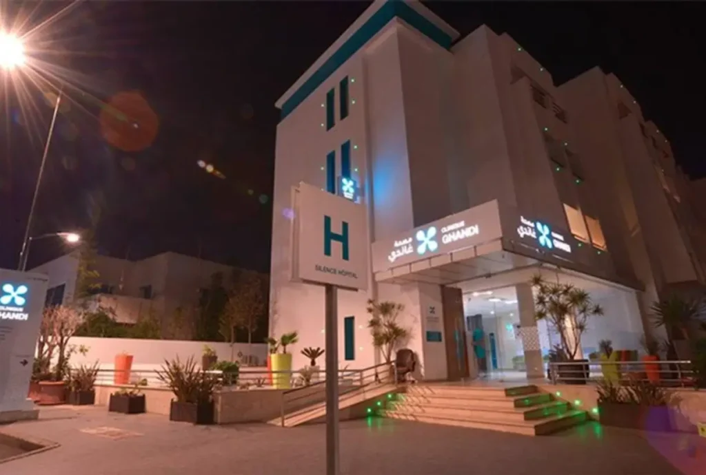 Façade éclairée d'une clinique privée moderne avec accès principal et signalétique hospitalière de nuit.